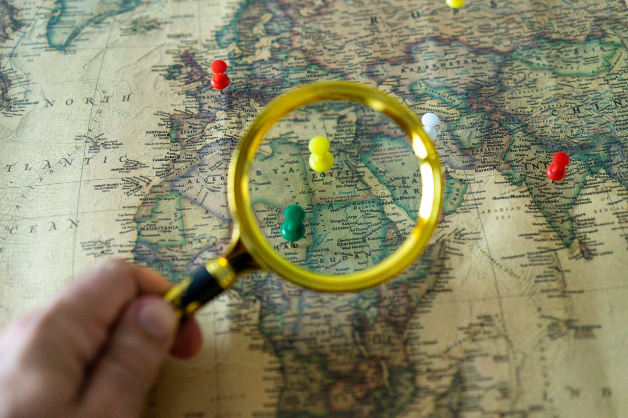 services-03 Close-up of a hand holding a magnifying glass over a world map with colorful pins indicating travel destinations.